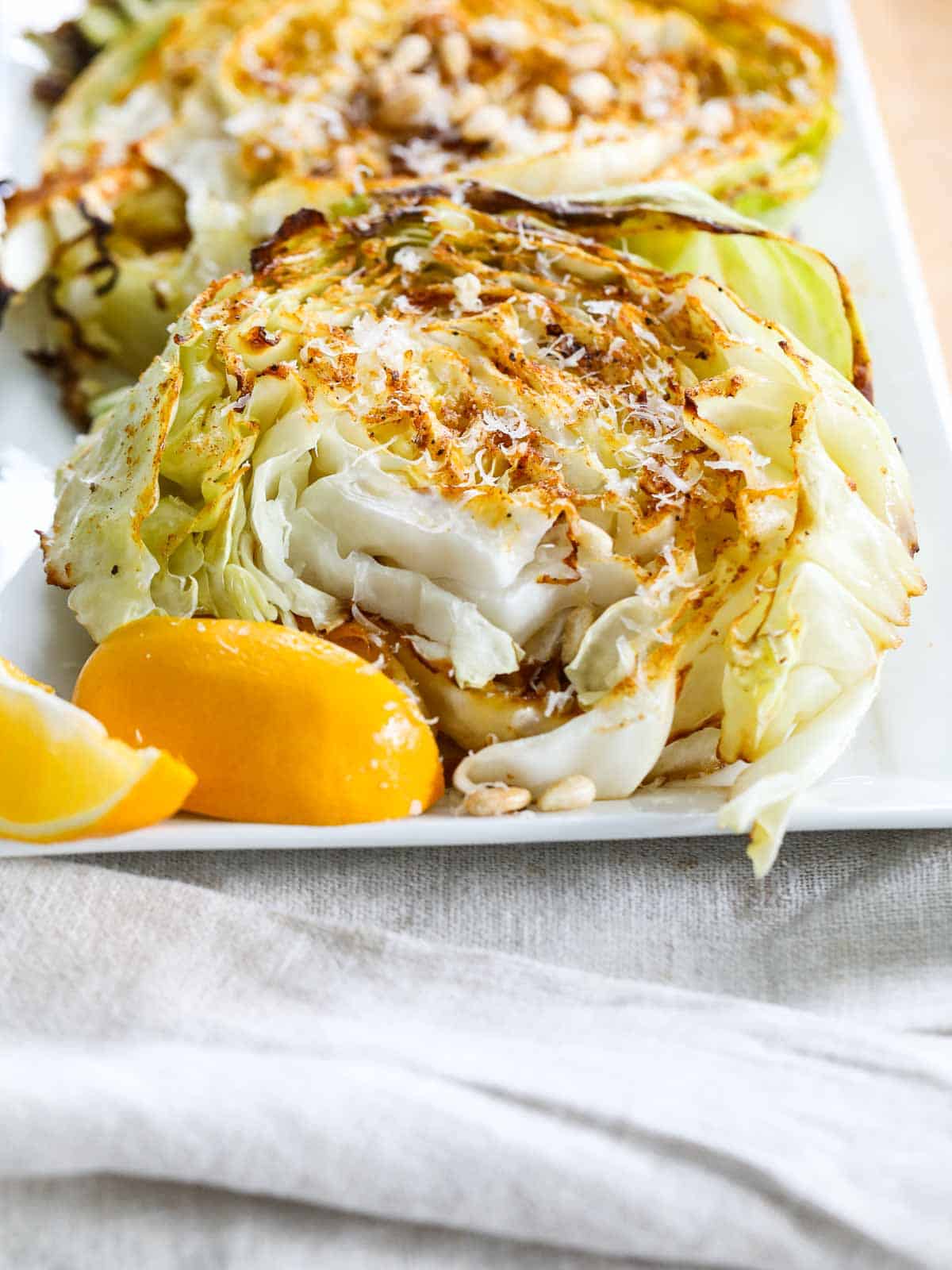 A white platter filled with cabbage steak, topped with shaved parmesan cheese and toasted pine nuts with lemon wedges.