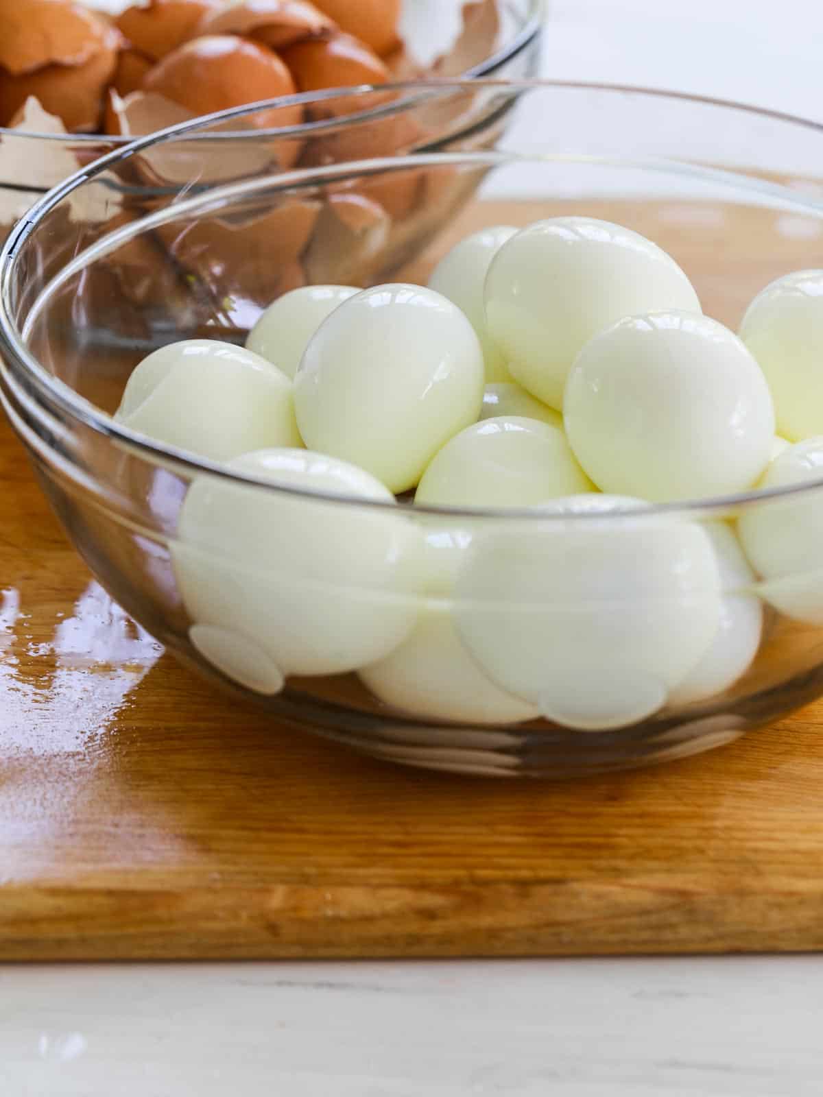 A large clear glass bowl filled with perfectly boiled eggs on a cutting board.