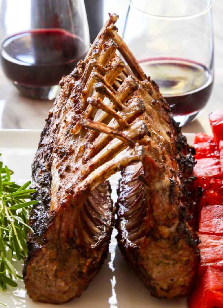 A beautiful grilled rack of lamb on a white platter with rosemary sprigs and watermelon skewers.