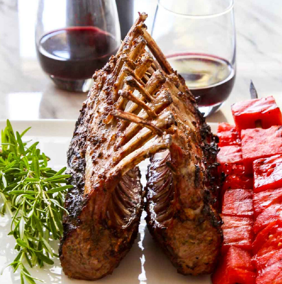 A beautiful grilled rack of lamb on a white platter with rosemary sprigs and watermelon skewers.