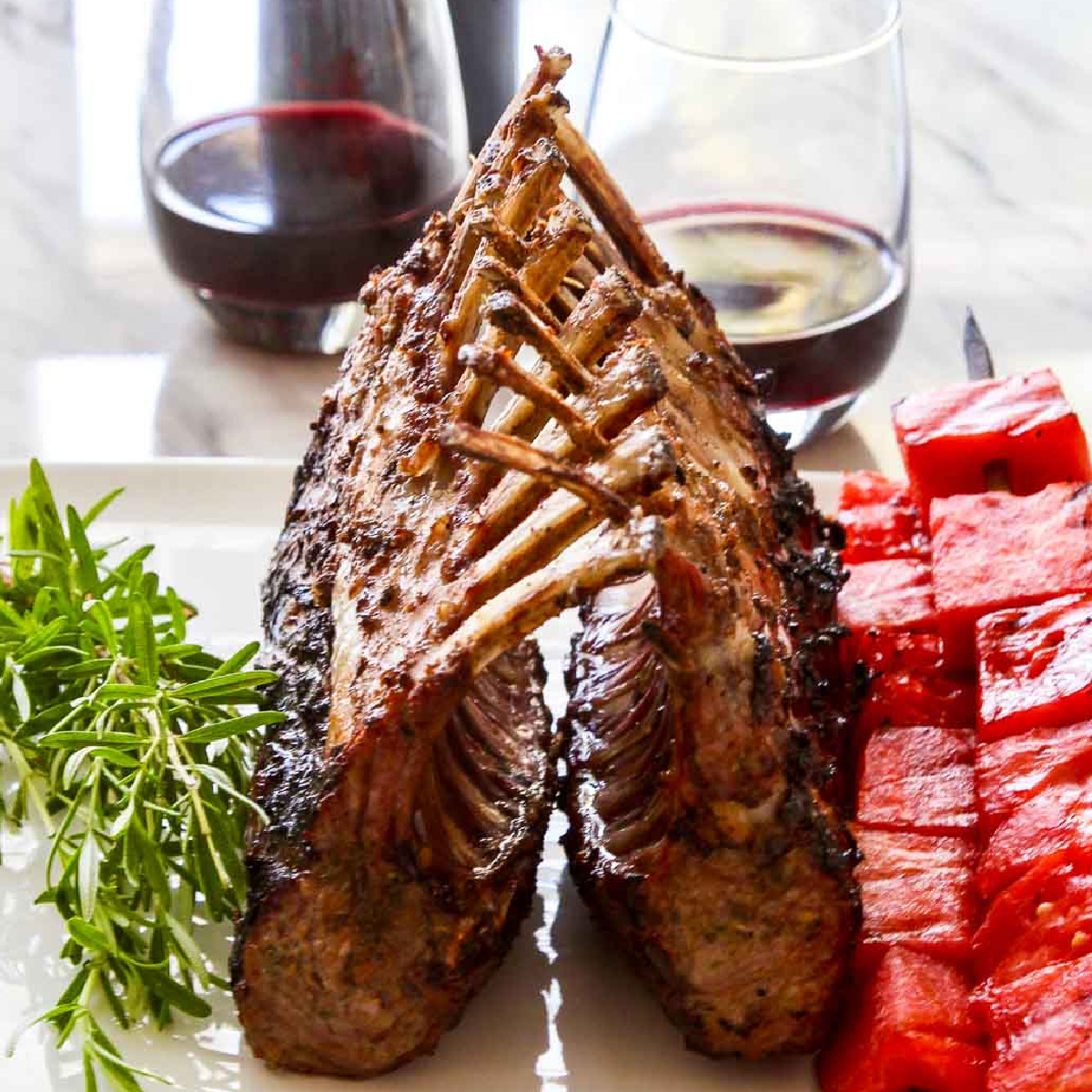 A beautiful grilled rack of lamb on a white platter with rosemary sprigs and watermelon skewers.