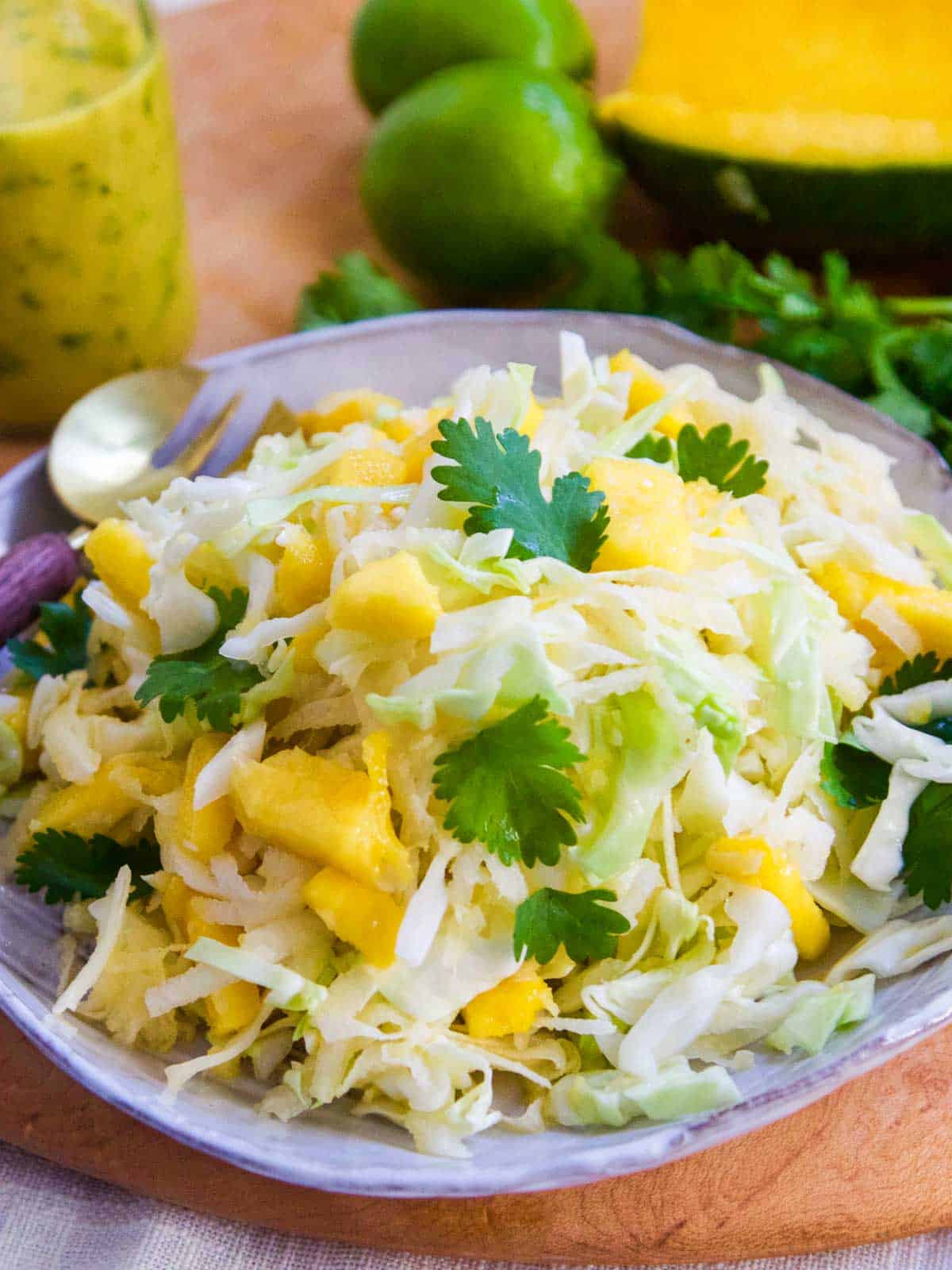 Mango slaw piled on a plate with a glass jar of dressing and limes in the background.