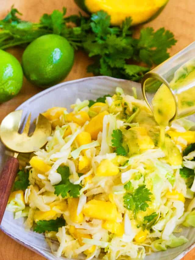 Pouring dressing on a full plate of mango slaw with a gold fork on edge of ceramic plate.