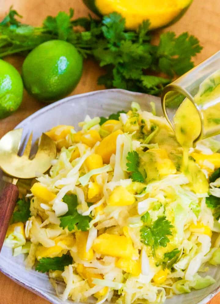 Pouring dressing on a full plate of mango slaw with a gold fork on edge of ceramic plate.