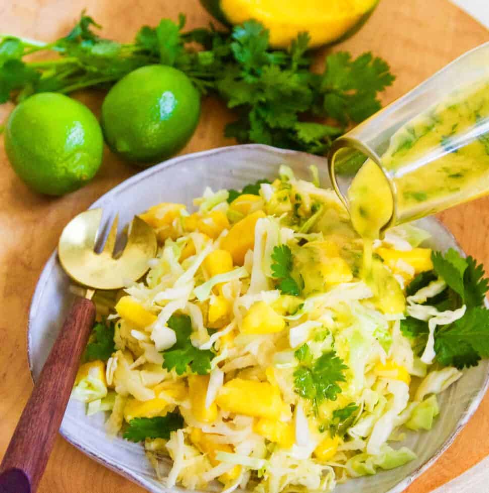 Pouring dressing on a full plate of mango slaw with a gold fork on edge of ceramic plate.