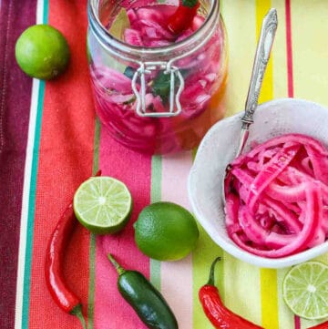 Mexican pickled red onions in a clamp jar and white bowl with chiles and limes on a colorful striped cloth.