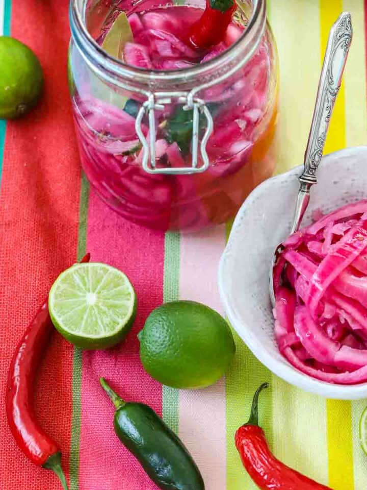 Mexican pickled red onions in a clamp jar and white bowl with chiles and limes on a colorful striped cloth.