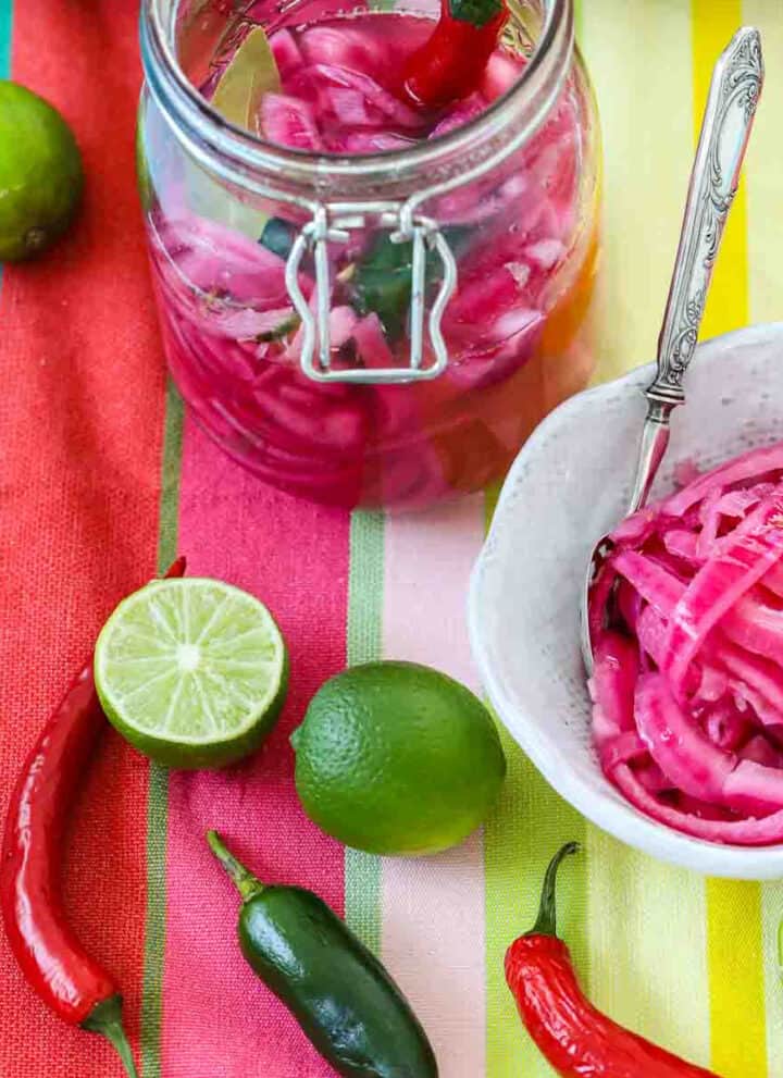 Mexican pickled red onions in a clamp jar and white bowl with chiles and limes on a colorful striped cloth.