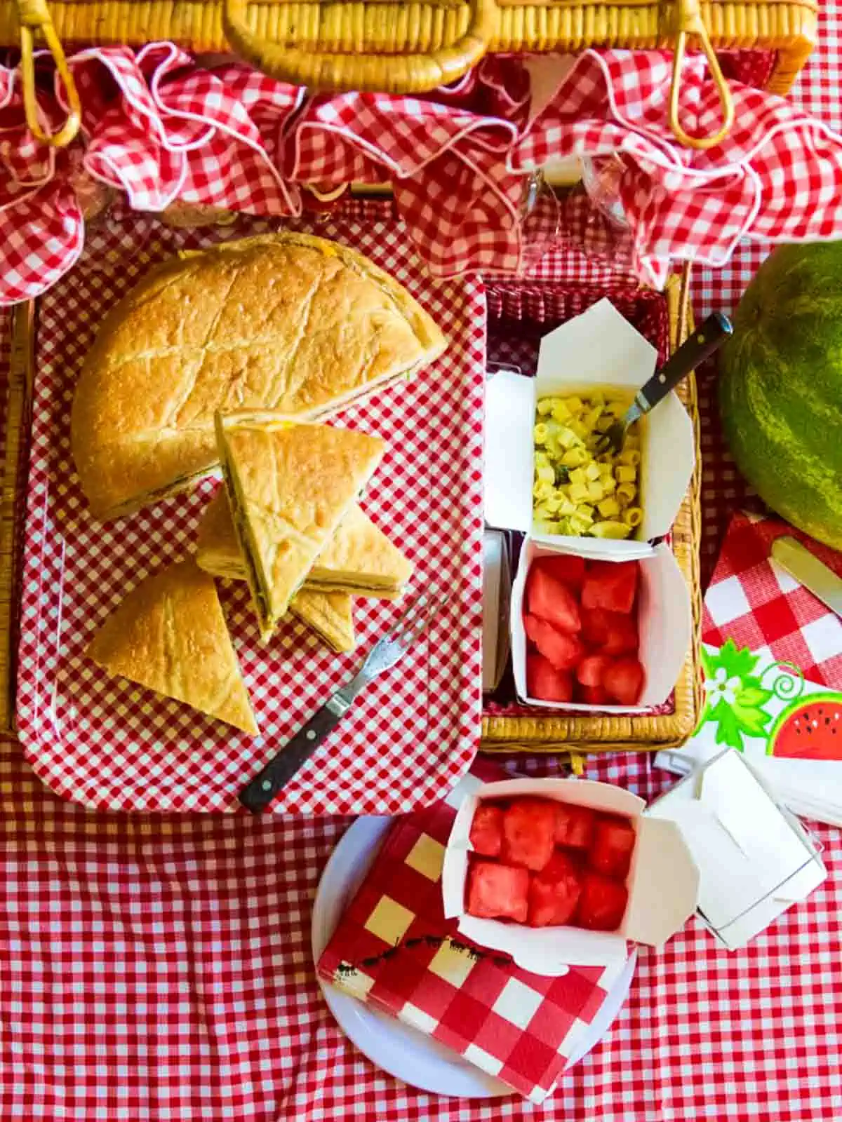 A red and white picnic basket with picnic sandwiches, watermelon and mac salad.