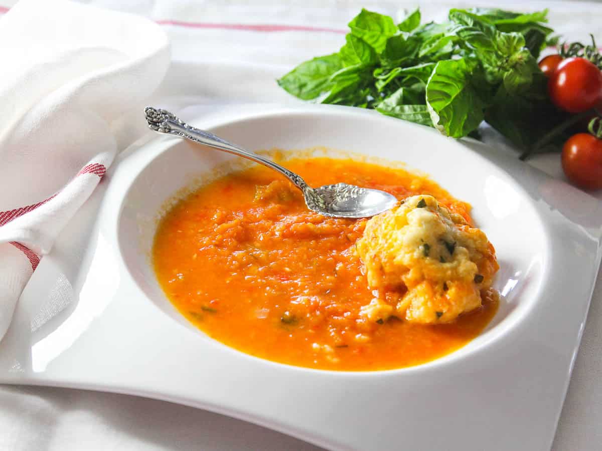 A large white bowl with tomato soup and a silver spoon with basil and tomatoes in the background.