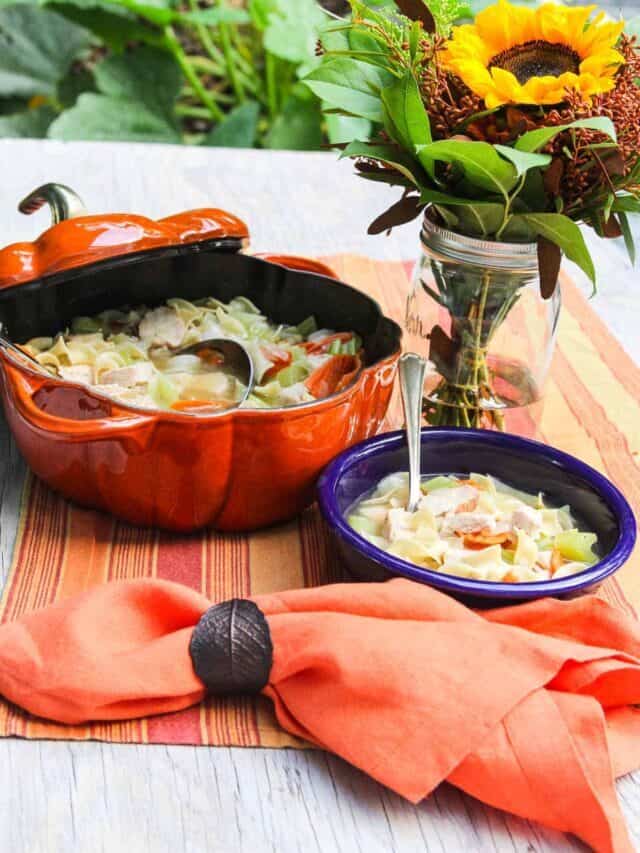 A large orange dutch oven filled with chicken noodle soup and a blue bowl with spoon and soup inside.