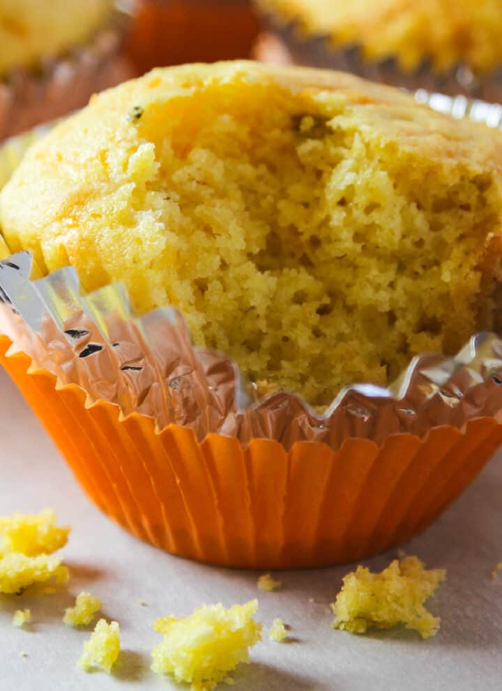 A cornbread muffin in an orange muffin tin paper with a piece out of it and crumbs on the table.