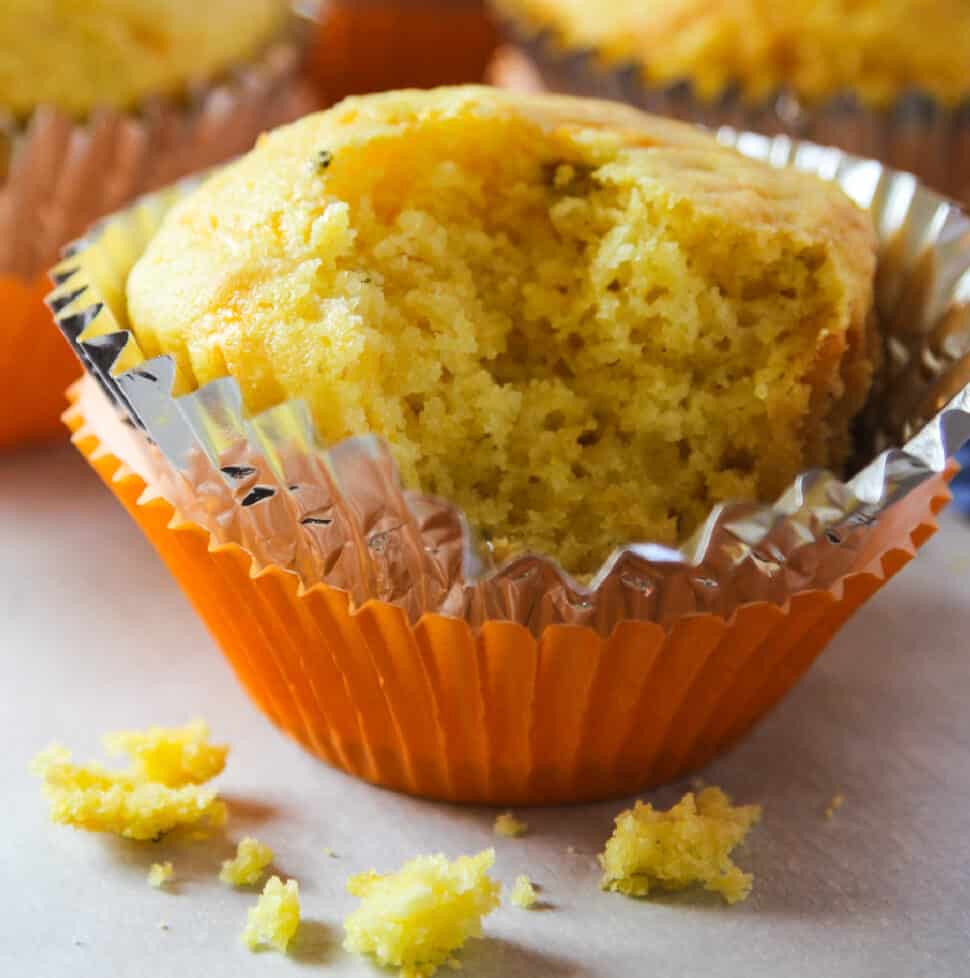 A cornbread muffin in an orange muffin tin paper with a piece out of it and crumbs on the table.