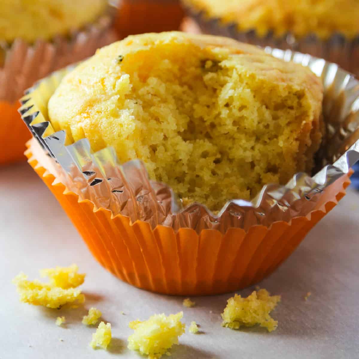 A cornbread muffin in an orange muffin tin paper with a piece out of it and crumbs on the table.