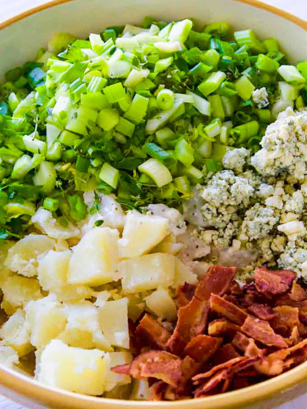A large mixing bowl with all the ingredients needed for a bacon potato salad recipe loaded inside before mixing those ingredients.