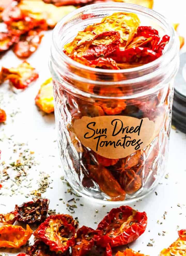 A small clear jar filled with sun dried tomatoes on a table with scattered dried tomatoes all around the table.
