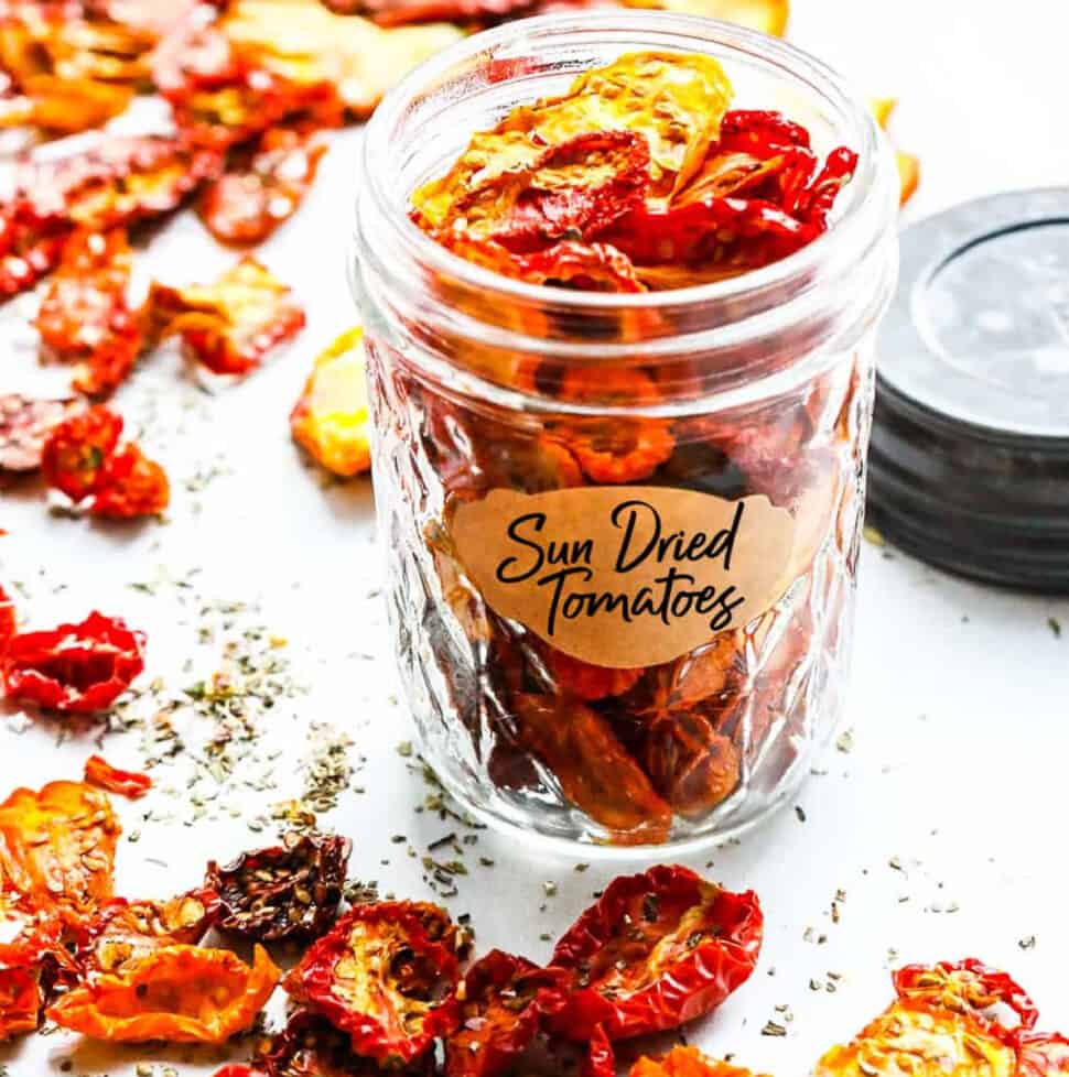 A small clear jar filled with sun dried tomatoes on a table with scattered dried tomatoes all around the table.