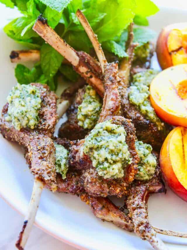 A white platter with grilled lamb chops topped with a mint pesto with grilled peaches on the side.