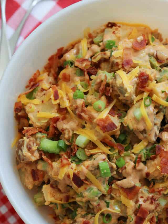 A large white bowl of BBQ potato salad garnished with cheddar cheese, bacon, and green onions on a red and white check gingham cloth.