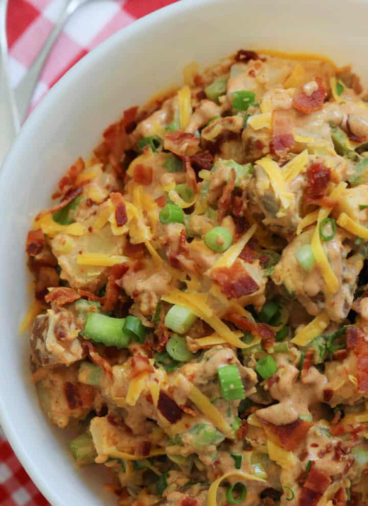 A large white bowl of BBQ potato salad garnished with cheddar cheese, bacon, and green onions on a red and white check gingham cloth.