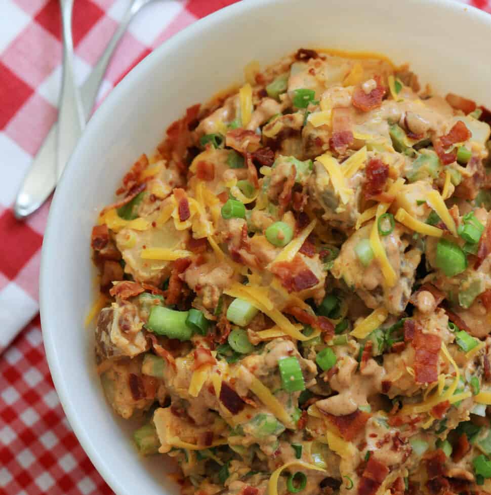 A large white bowl of BBQ potato salad garnished with cheddar cheese, bacon, and green onions on a red and white check gingham cloth.