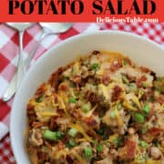 A large white bowl of BBQ potato salad garnished with cheddar cheese, bacon, and green onions on a red and white check gingham cloth.