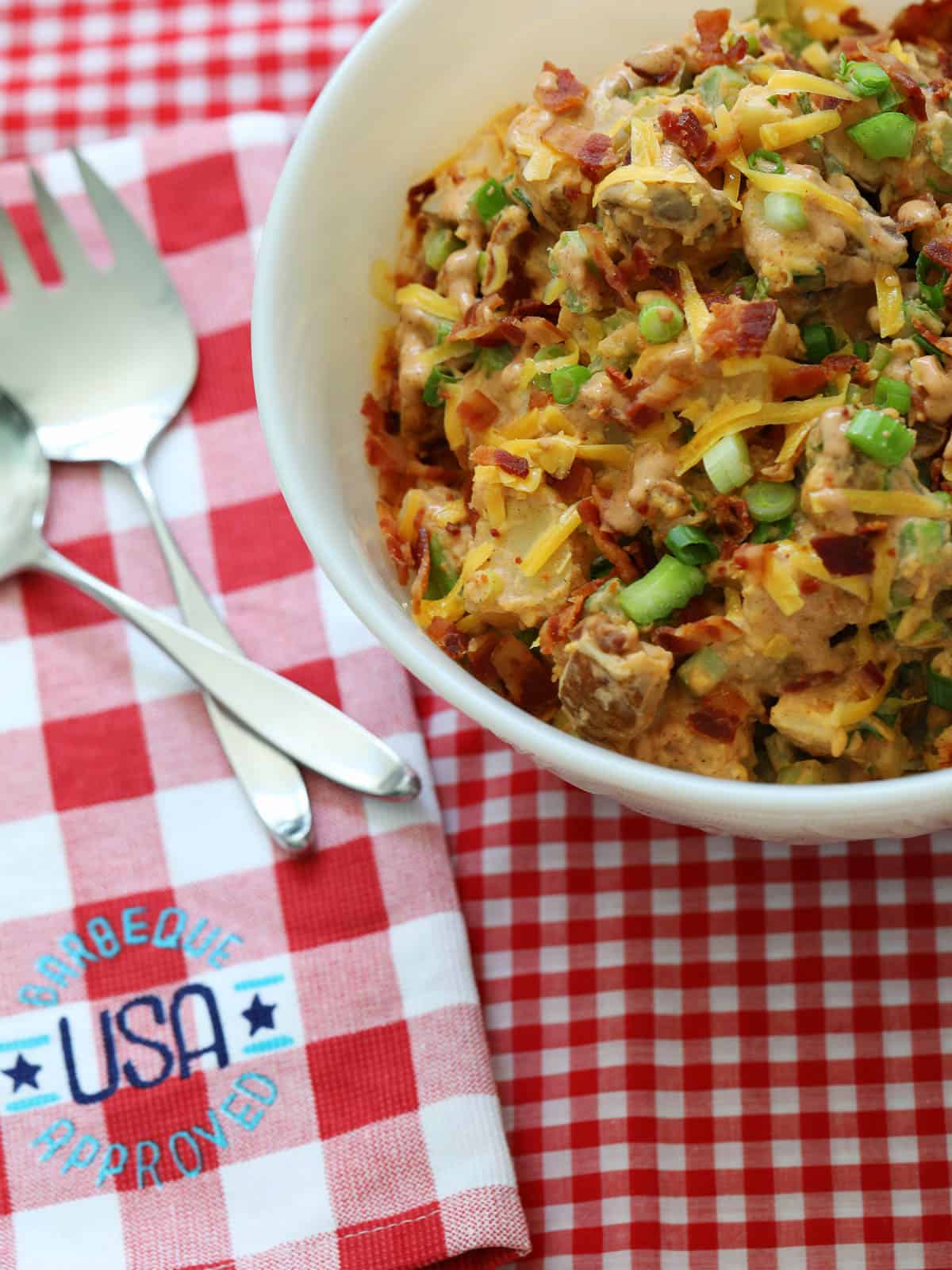 A large white bowl of BBQ potato salad garnished with cheddar cheese, bacon, and green onions on a red and white check gingham cloth.