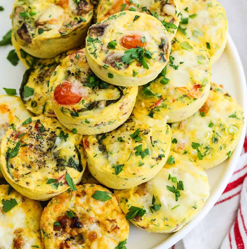 Small round breakfast egg muffins stacked on a white cake plate for a brunch or breakfast filled with different flavors.