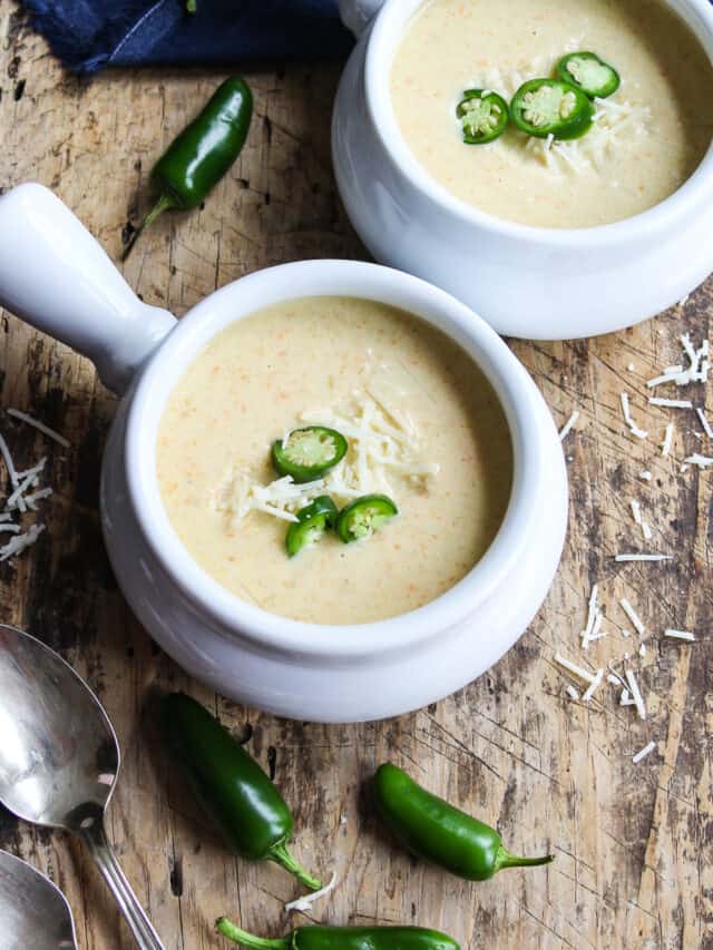 Two bowls of creamy jalapeno popper soup in white handled soup bowls garnished with thin sliced jalapenos and cheese.