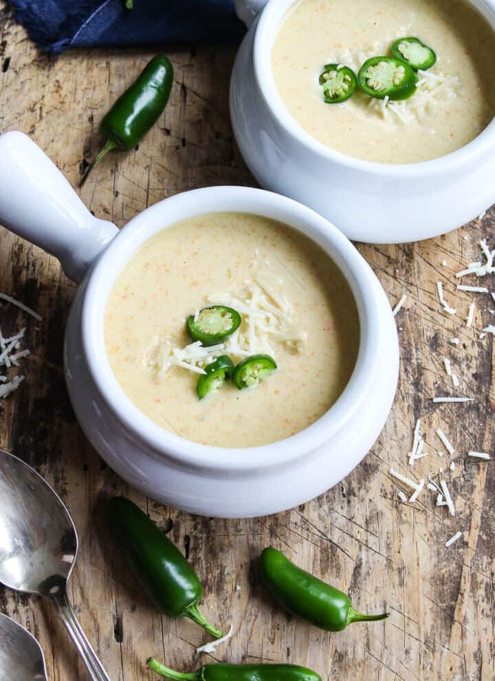 Two bowls of creamy jalapeno popper soup in white handled soup bowls garnished with thin sliced jalapenos and cheese.