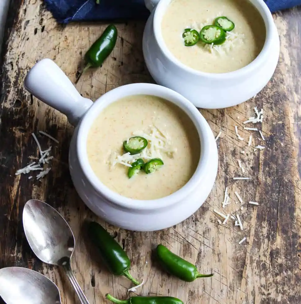 Two bowls of creamy jalapeno popper soup in white handled soup bowls garnished with thin sliced jalapenos and cheese.