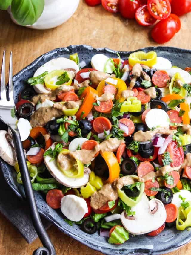 A plate full of pizza salad topped with mini pepperoni, mushrooms, mozzarella balls, pepperoncinis, and black olives with a artistic black fork on the edge of the plate.