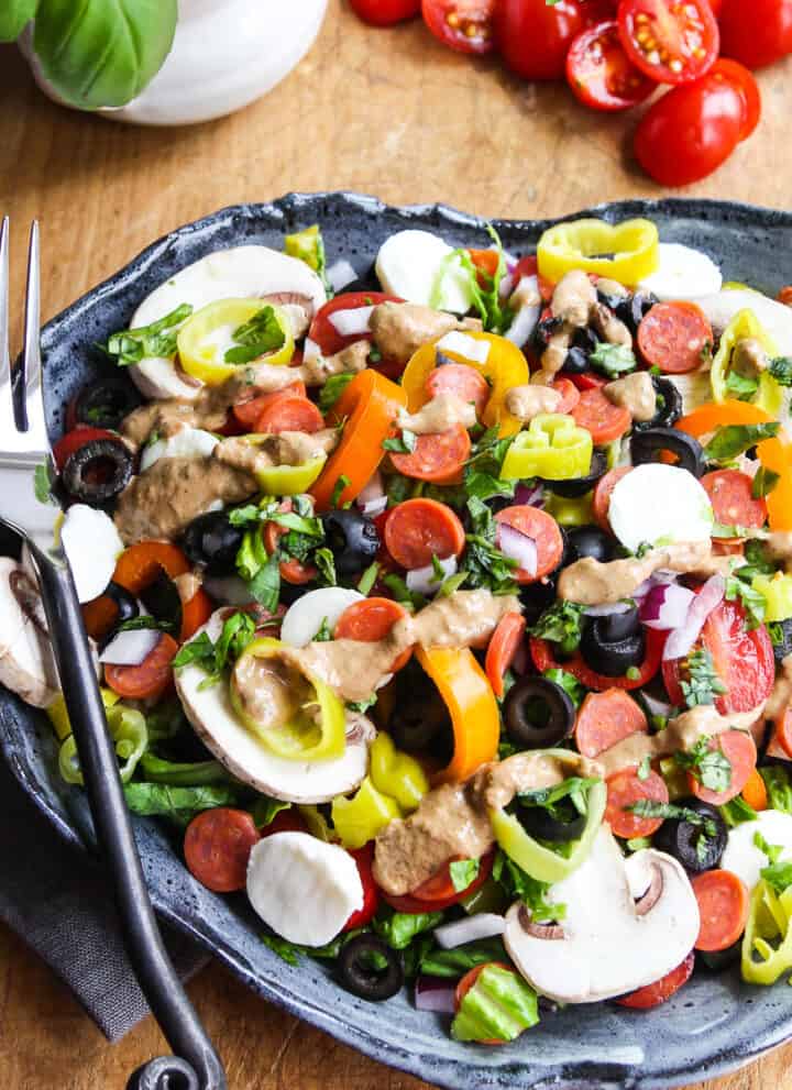 A plate full of pizza salad topped with mini pepperoni, mushrooms, mozzarella balls, pepperoncinis, and black olives with a artistic black fork on the edge of the plate.