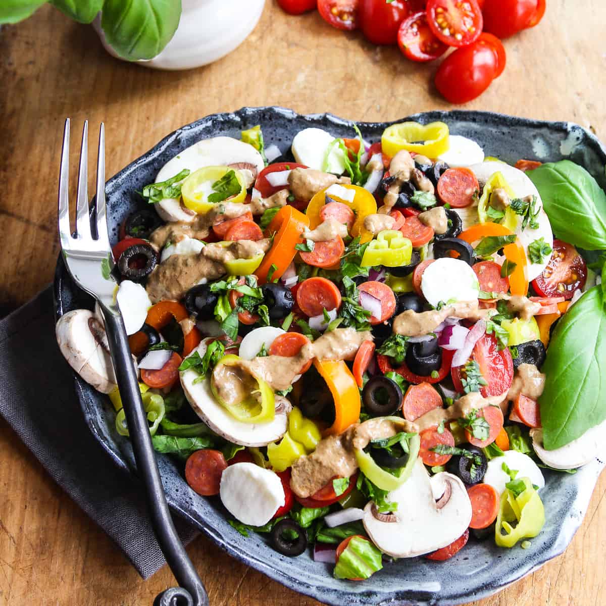 A plate full of pizza salad topped with mini pepperoni, mushrooms, mozzarella balls, pepperoncinis, and black olives with a artistic black fork on the edge of the plate.