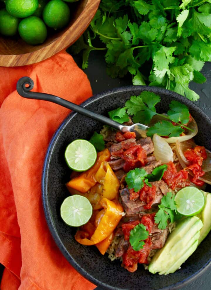 A table with a black bowl filled with shredded beef on rice with onions and bell peppers garnished with limes, avocado, and cilantro.