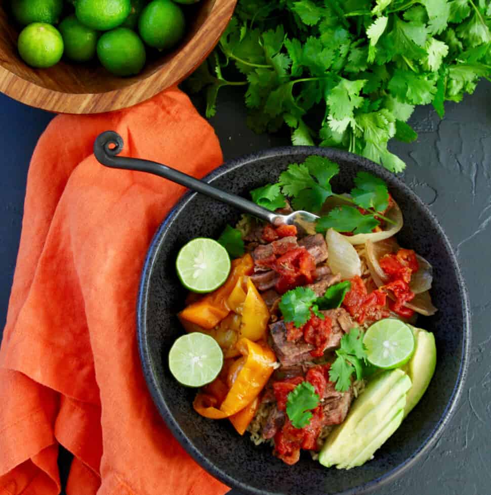 A table with a black bowl filled with shredded beef on rice with onions and bell peppers garnished with limes, avocado, and cilantro.