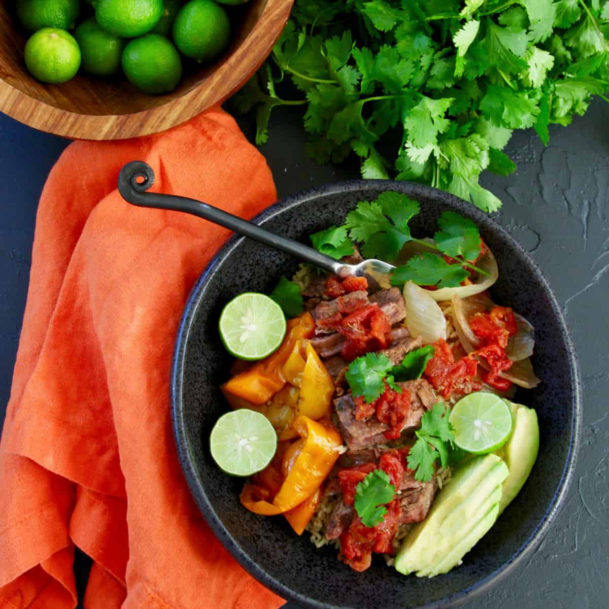 A table with a black bowl filled with shredded beef on rice with onions and bell peppers garnished with limes, avocado, and cilantro.