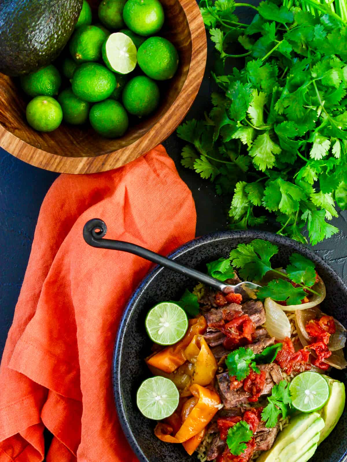 A wood bowl of limes with slow cooker shredded beef topped with cilantro, avocado, and limes.