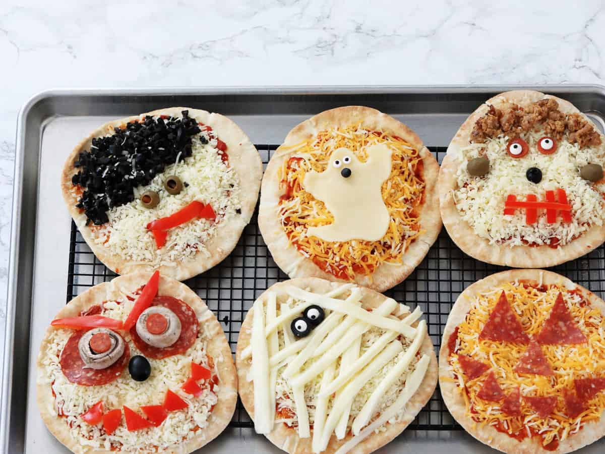 Six Halloween pizzas on a baking rack decorated to look like ghosts, pumpkins, dracula, frankenstein, and monsters.