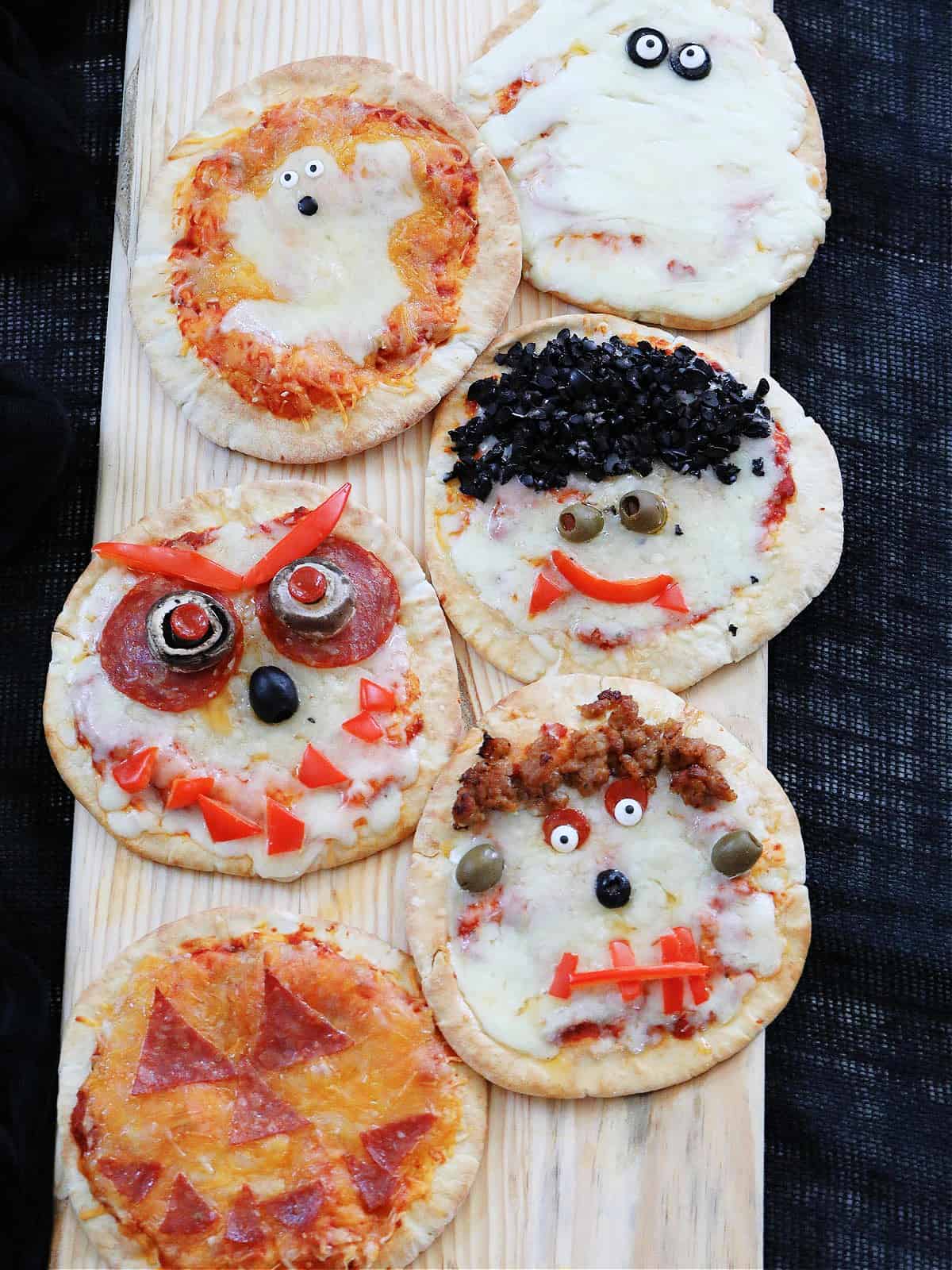 Six baked Halloween pizzas on a board decorated to look like ghosts, pumpkins, dracula, frankenstein, and monsters.