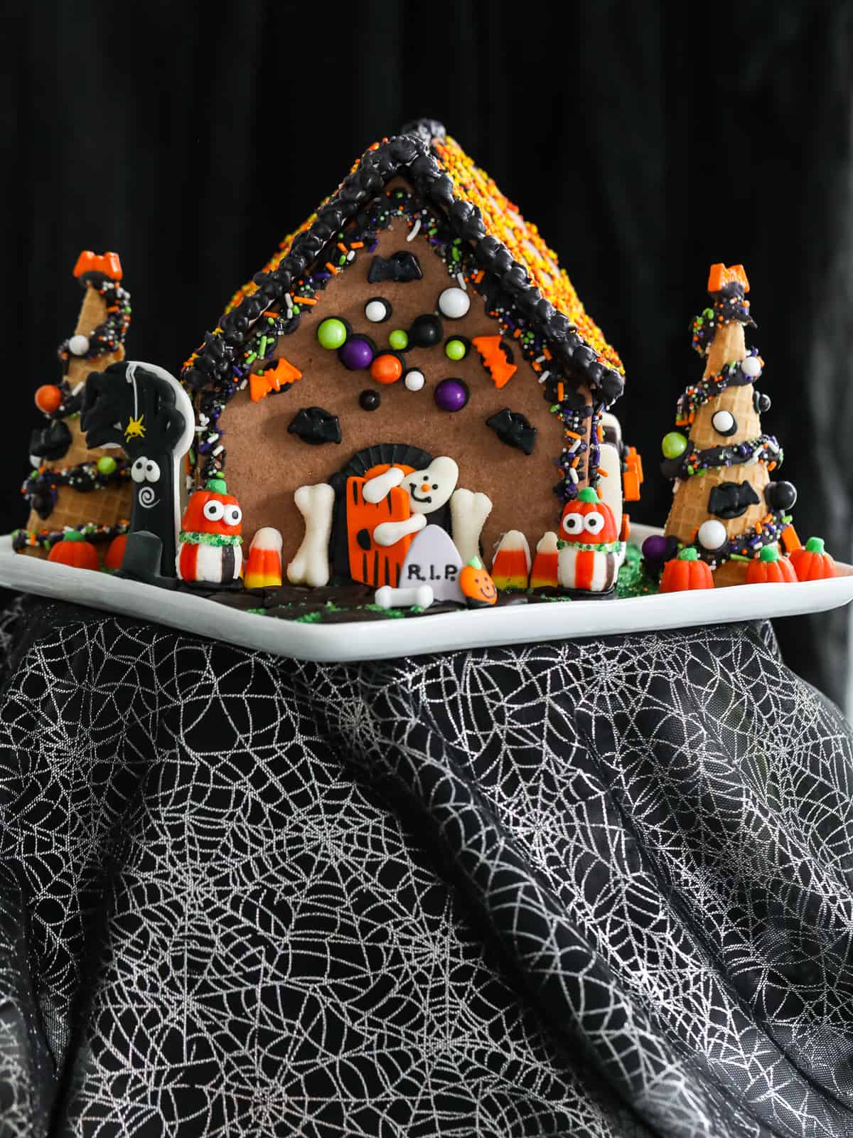 A Halloween gingerbread house all decorated with bright candy on a white plate at a Halloween party.