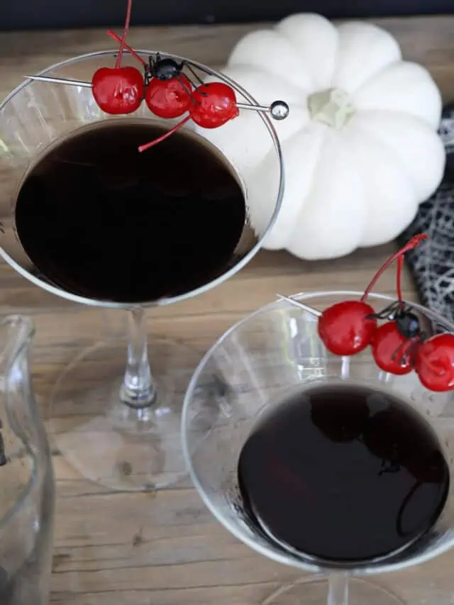 Two Halloween black drinks make with black vodka garnished with 3 red maraschino cherries and a black spider.