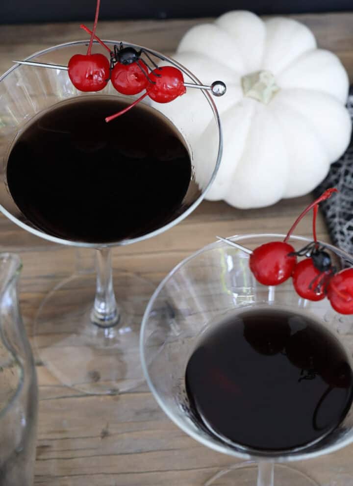 Two Halloween black drinks make with black vodka garnished with 3 red maraschino cherries and a black spider.