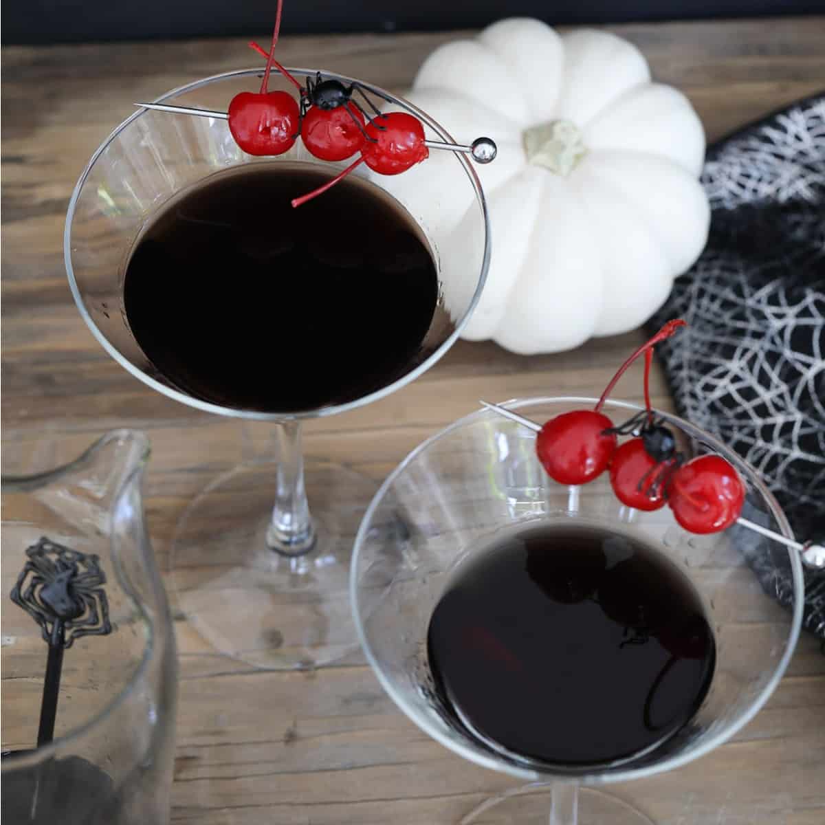 Two Halloween black drinks make with black vodka garnished with 3 red maraschino cherries and a black spider.