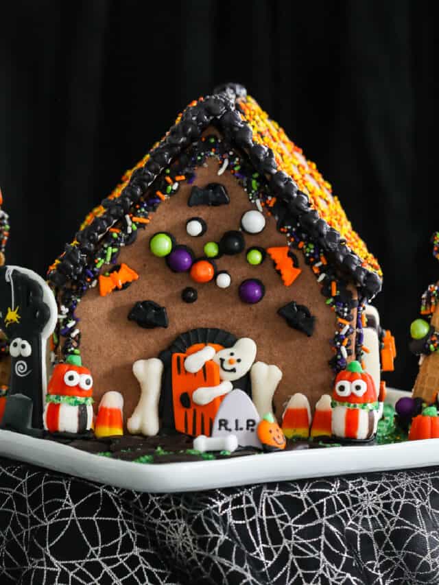 A Halloween gingerbread house decorated with royal icing and candy in orange, black, and white candy with a ghost opening the front door.