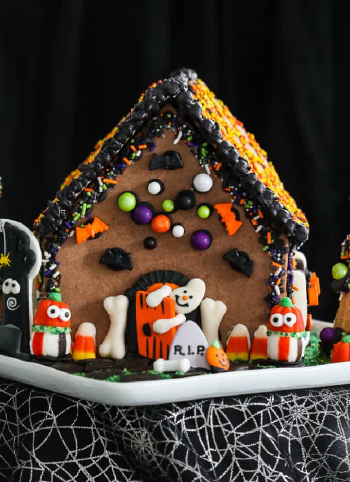 A Halloween gingerbread house decorated with royal icing and candy in orange, black, and white candy with a ghost opening the front door.