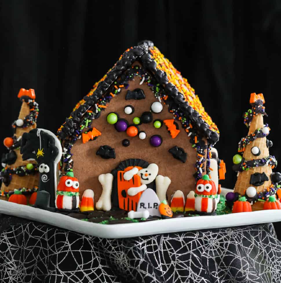 A Halloween gingerbread house decorated with royal icing and candy in orange, black, and white candy with a ghost opening the front door.