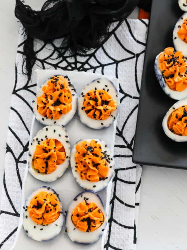 A white ceramic plate with six Halloween deviled eggs on a black and white spider tablecloth at a Halloween party.