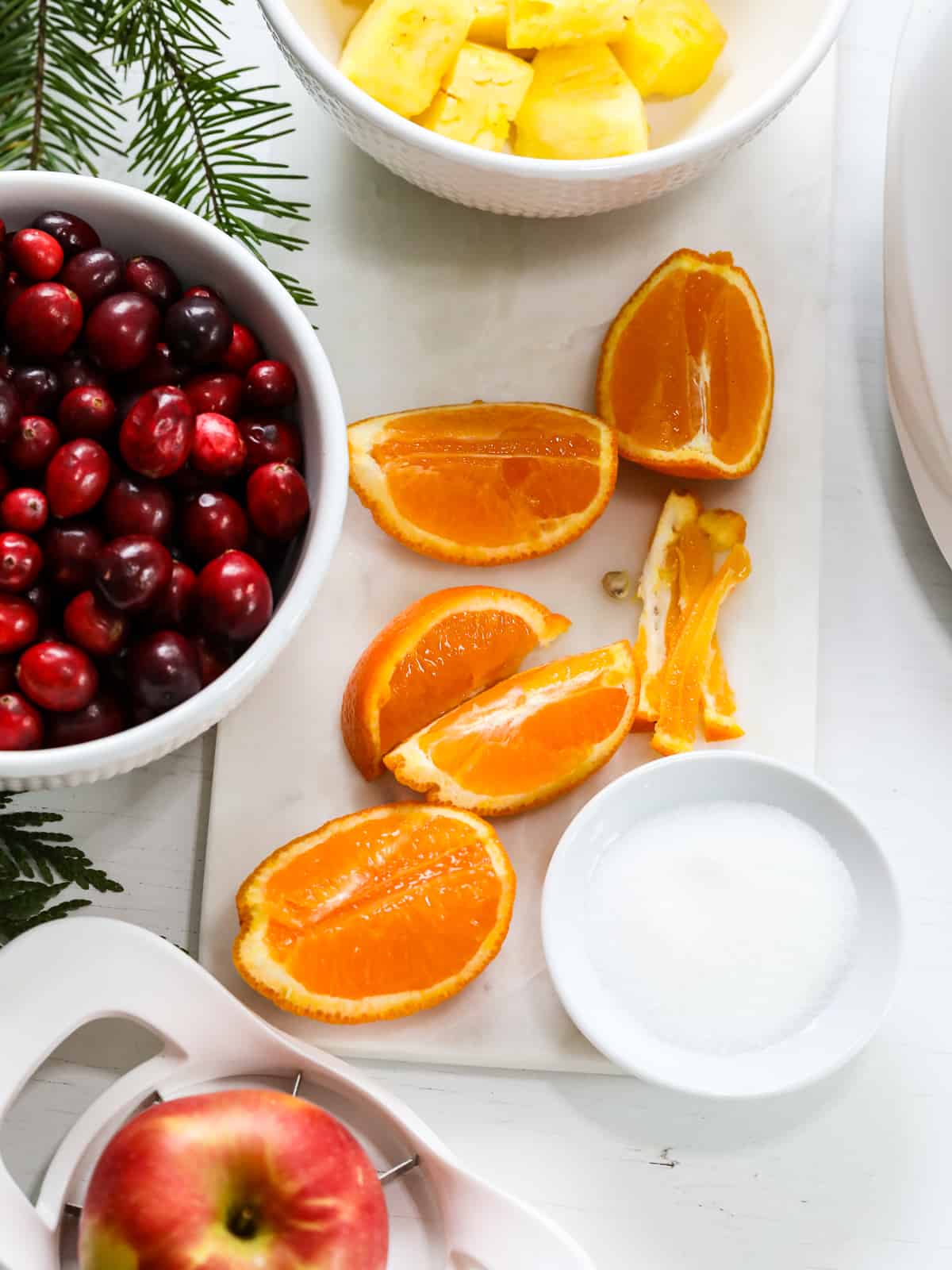 The ingredients to make cranberry relish on a white table including cranberries, oranges, apple , and sugar.