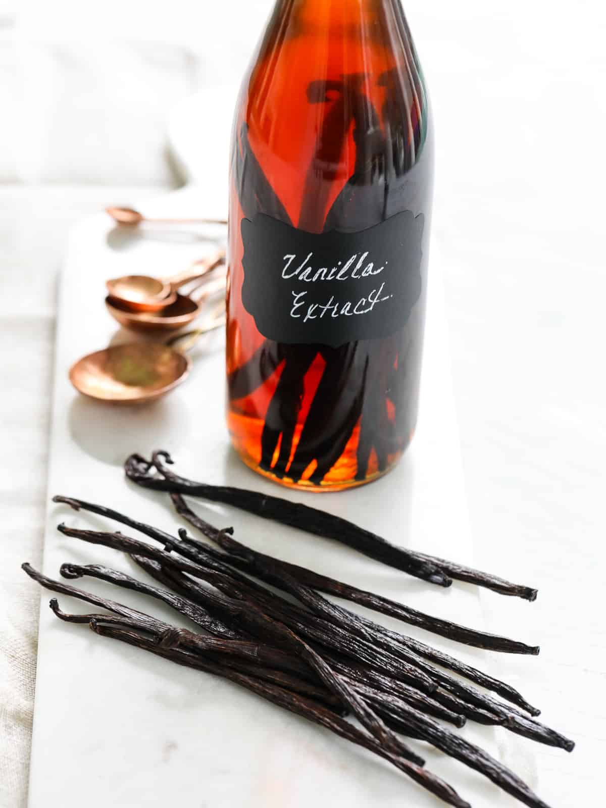 A tall glass bottle filled with vanilla extract and you can see the vanilla beans inside on a table with copper measuring spoons.
